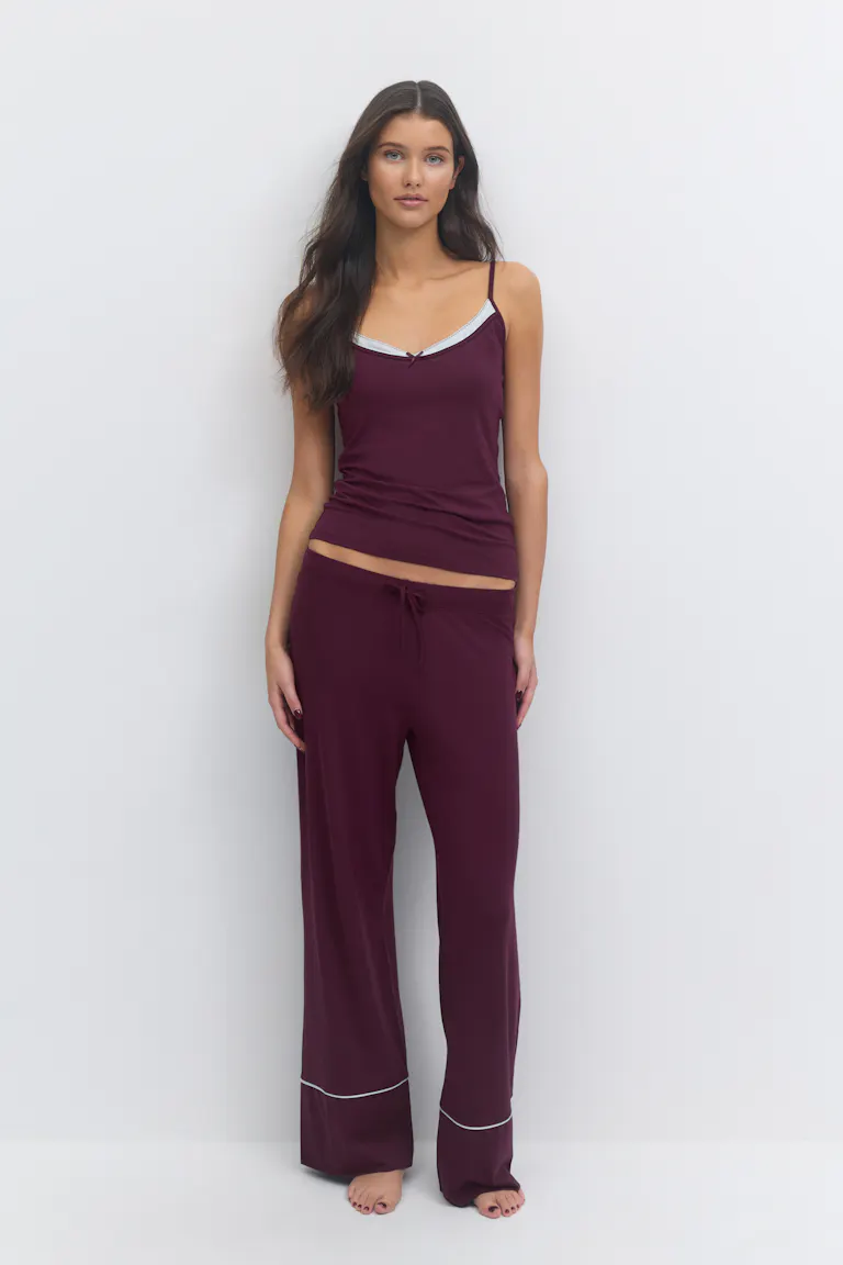 Syvä viininpunainen camisole, jossa on ohuet olkaimet, valkoisella reunustettu V-kaula-aukko ja pieni keskellä oleva rusetti, yhdistettynä yhteensopiviin leveälahkeisiin leveälahkeisiin oleskeluhousuihin, joissa on valkoinen putkitus reunojen yläpuolella.