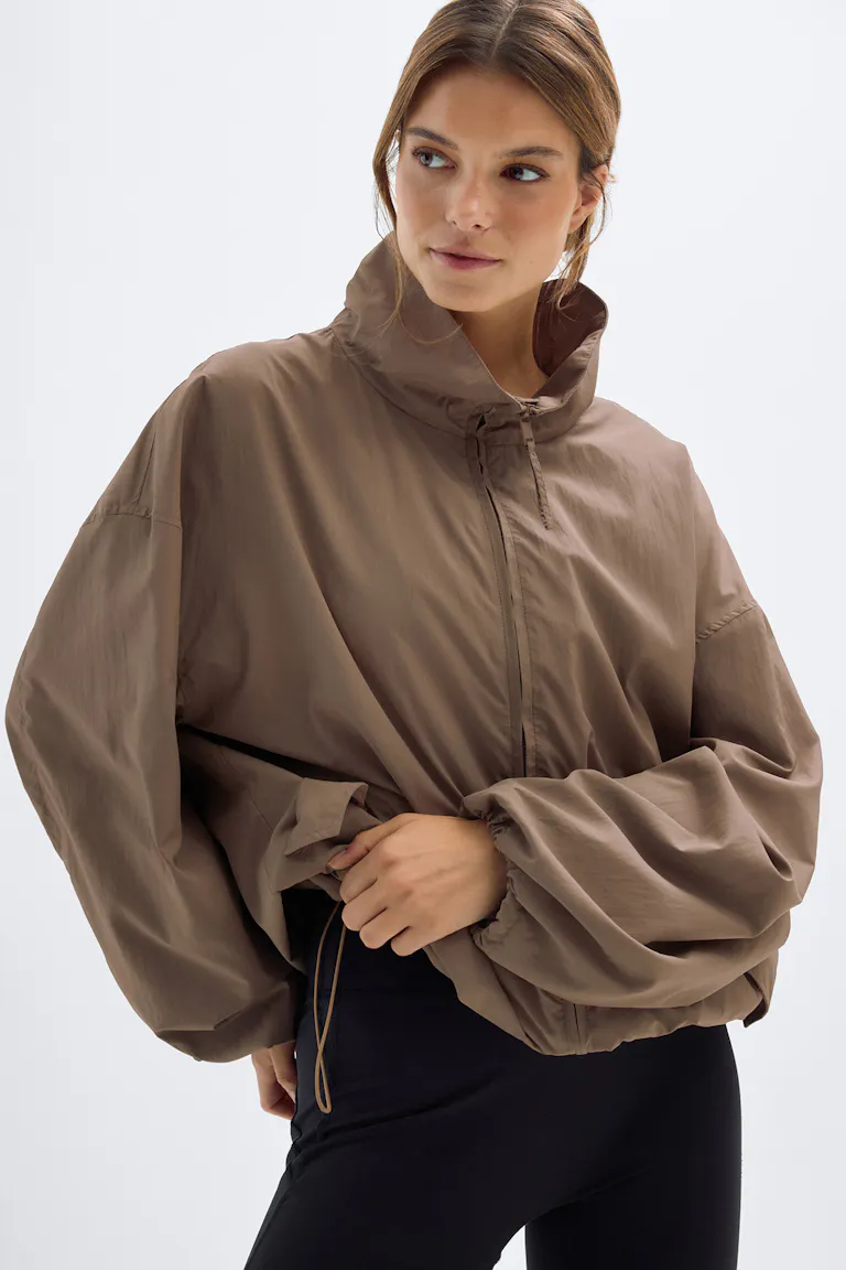 Taupe zip-front lightweight windbreaker with a high stand collar, flap pockets, elasticated cuffs and adjustable drawcord hem, paired with black leggings.