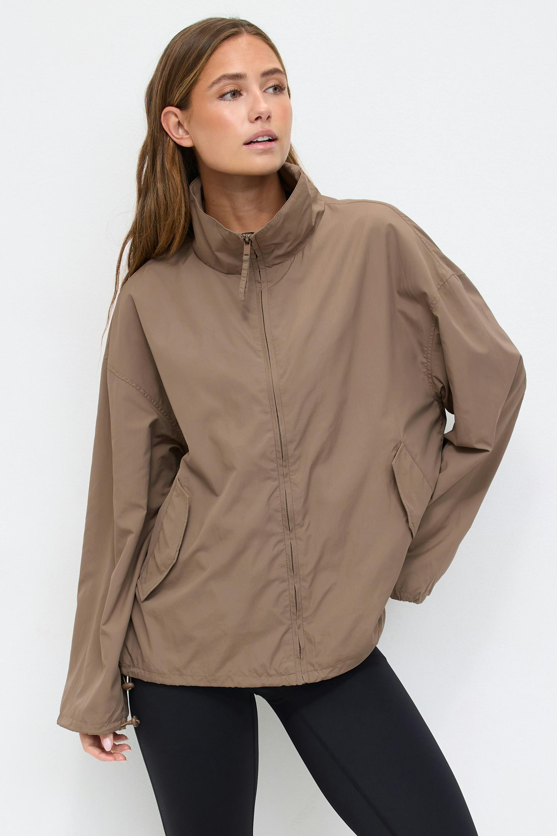 Taupe zip-front lightweight windbreaker with a high stand collar, flap pockets, elasticated cuffs and adjustable drawcord hem, paired with black leggings.
