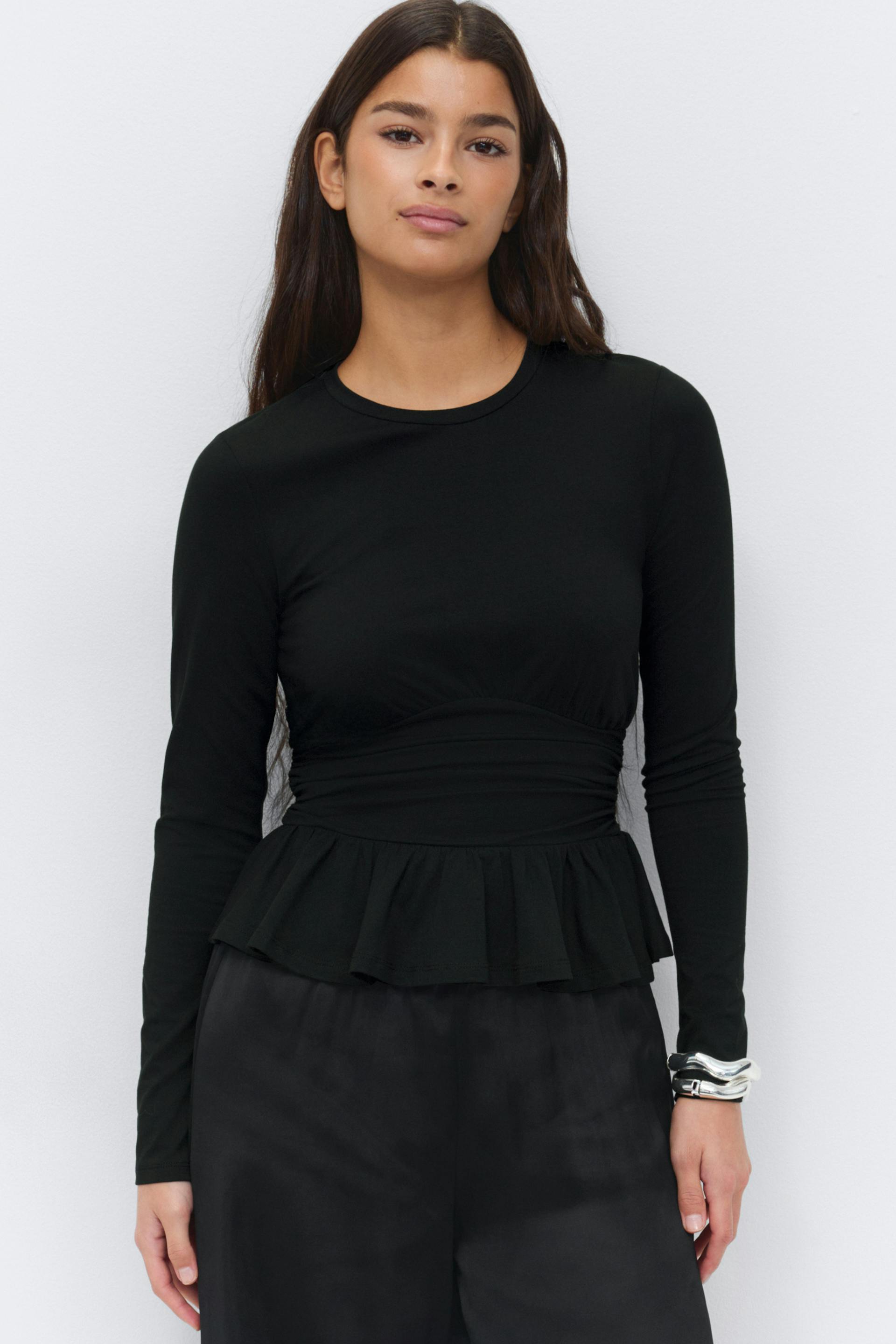 Black long-sleeve fitted peplum top with crew neckline and ruched waist, paired with black satin wide-leg trousers and silver cuff bracelets.