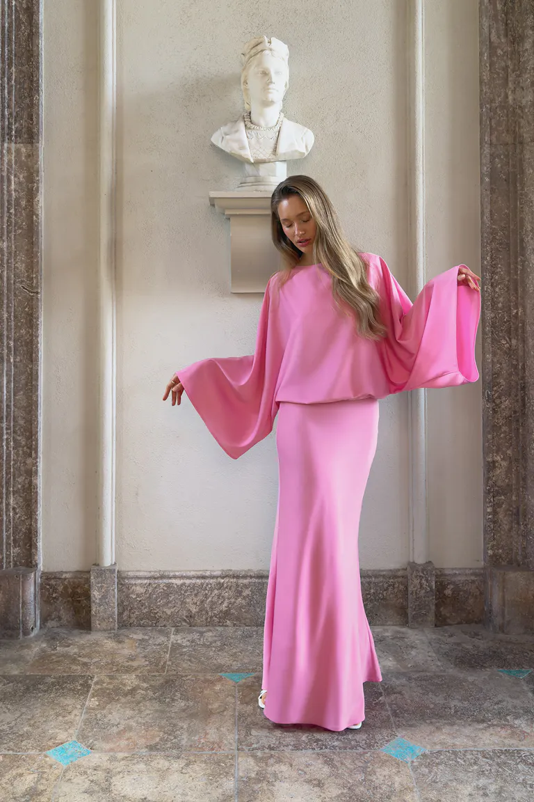 Bodenlanges rosafarbenes Kleid mit Blouson-Mieder, weiten drapierten Dolman-Ärmeln, tailliertem Rock und weißen offenen Absätzen.