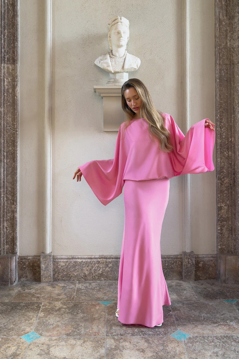 Floor-length pink gown with a blouson bodice, wide draped dolman sleeves, fitted skirt, and white open-toe heels.