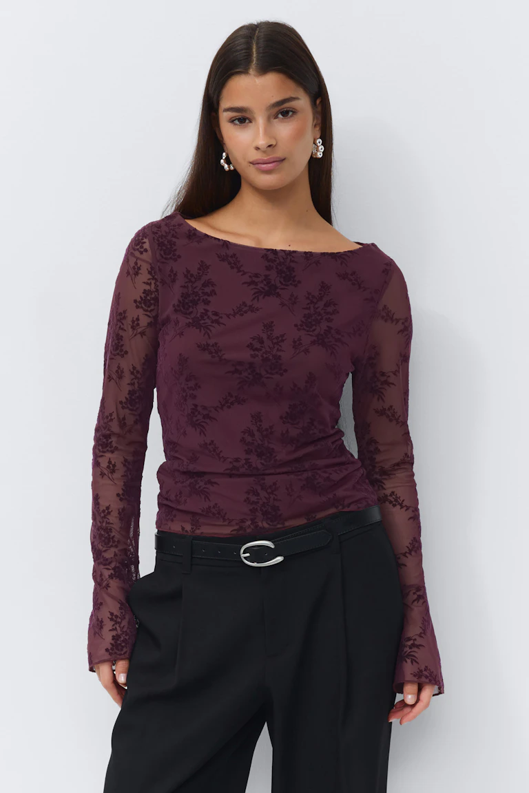 Sheer long-sleeve burgundy floral burnout mesh top with a boat neckline, tucked into high‑waisted black tailored trousers with a black leather belt featuring a silver oval buckle; paired with pearl cluster earrings.