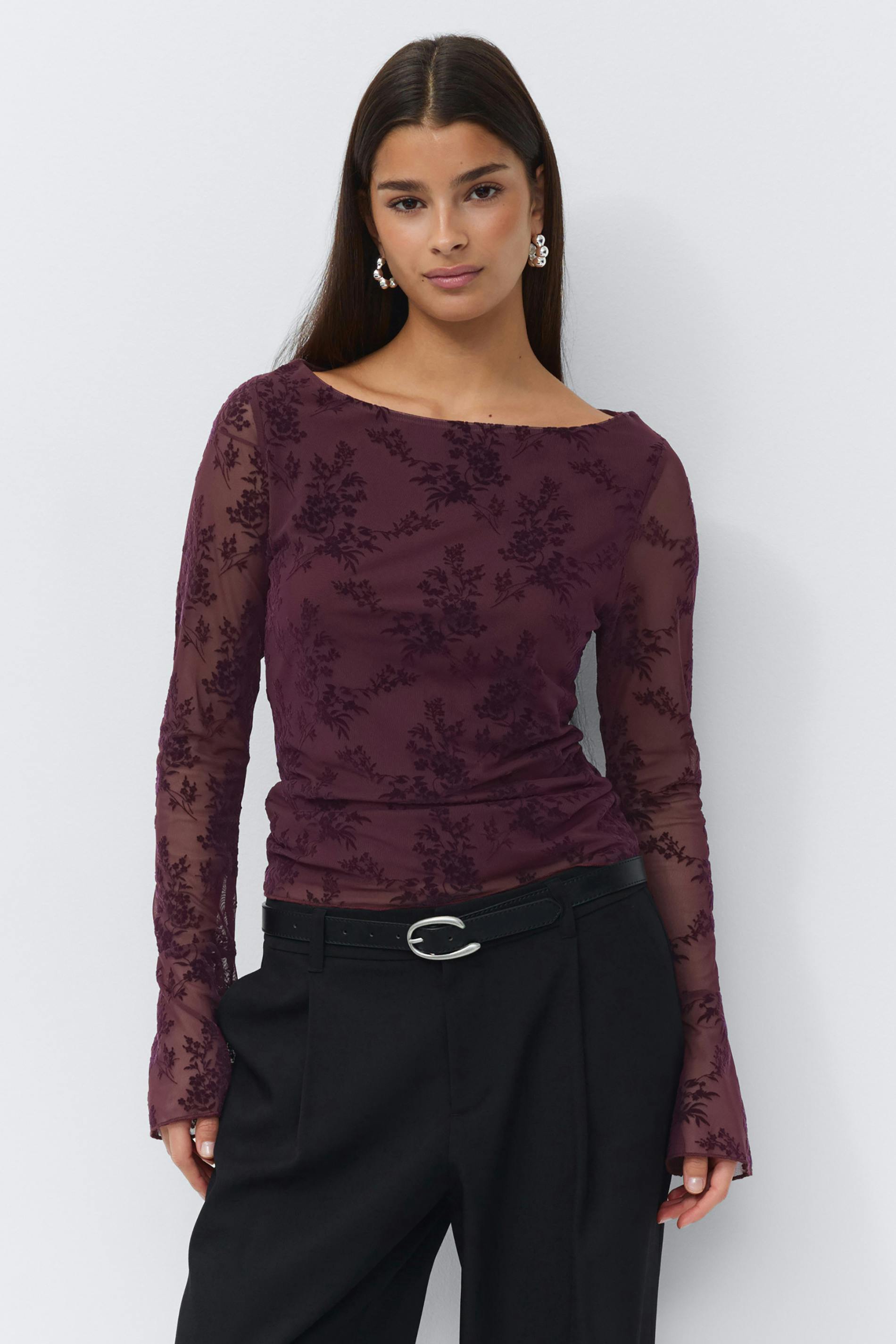 Sheer long-sleeve burgundy floral burnout mesh top with a boat neckline, tucked into high‑waisted black tailored trousers with a black leather belt featuring a silver oval buckle; paired with pearl cluster earrings.