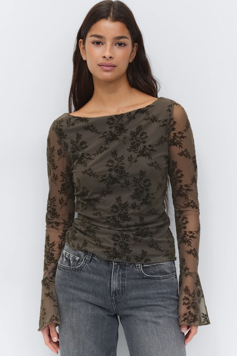 Top en maille transparente à manches longues olive foncé avec motif floral floqué noir, encolure bateau, ruchés subtils sur les côtés et poignets évasés, associé à un jean taille haute gris délavé.