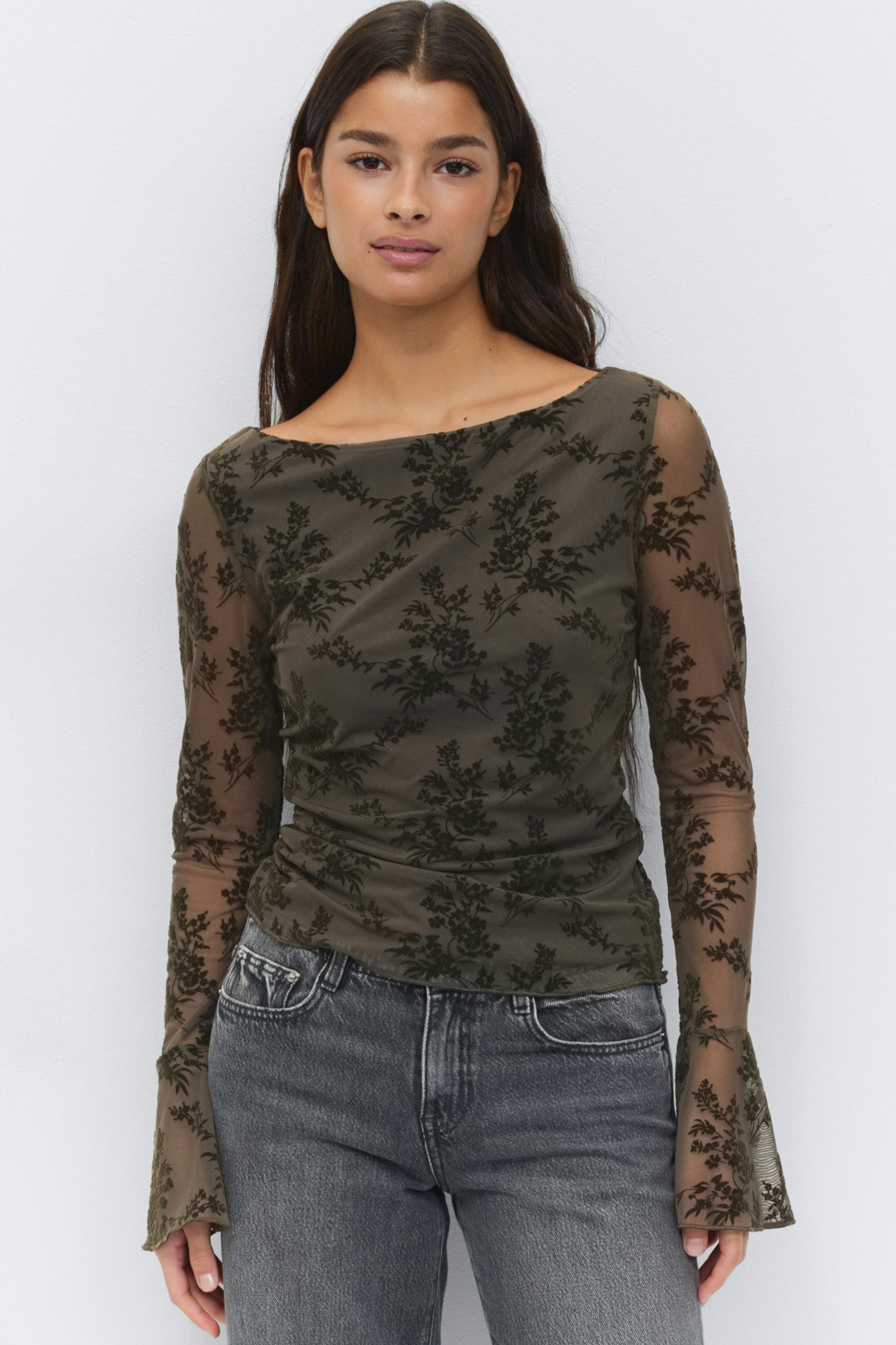 Dark olive sheer long-sleeve mesh top with black flocked floral pattern, boat neckline, subtle side ruching and flared cuffs, paired with faded grey high‑waist jeans.