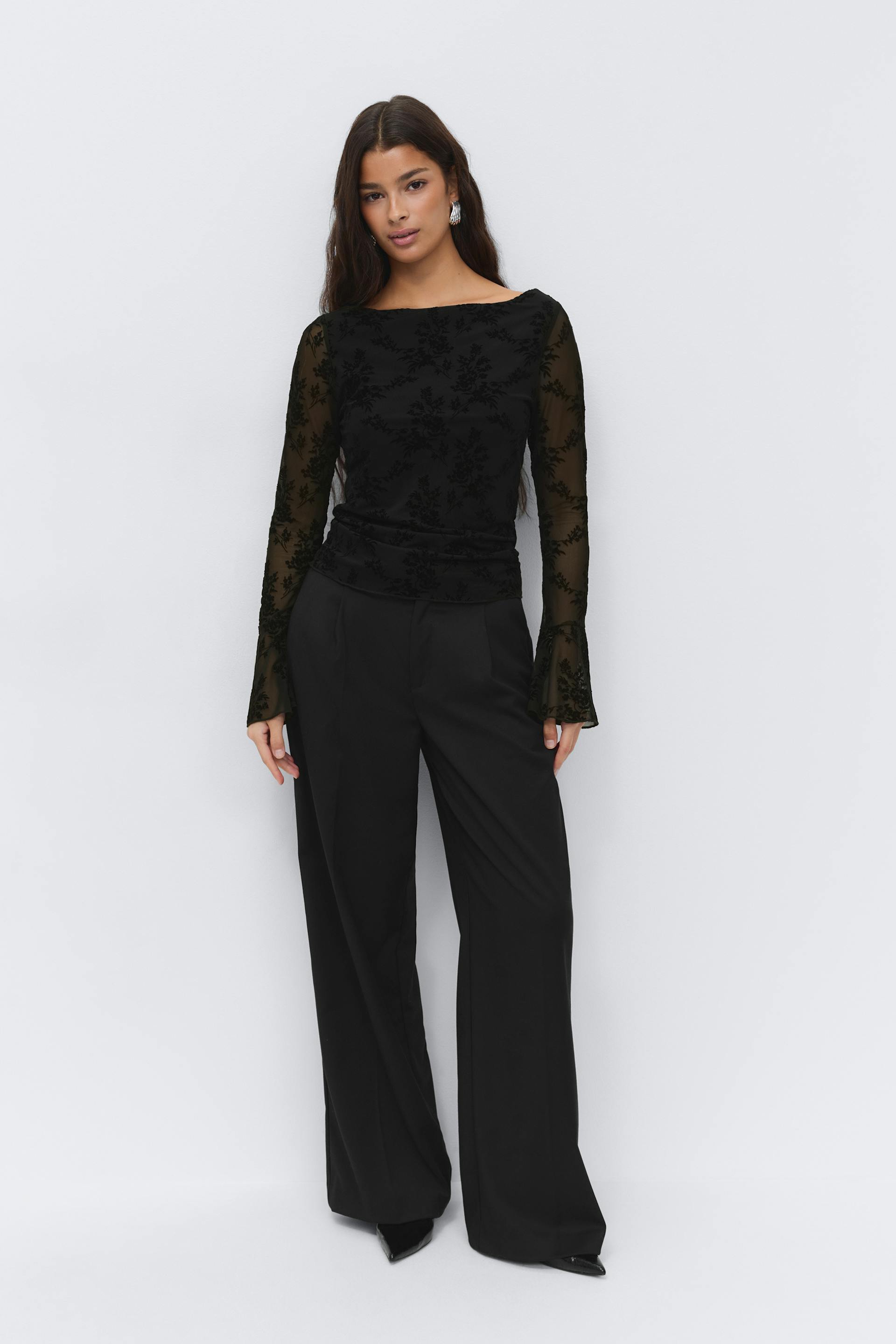 Black sheer long-sleeve boat-neck top with flocked floral pattern and flared cuffs, paired with high‑waisted wide‑leg black trousers and pointed black pumps.