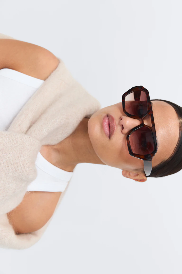Weißes geripptes Tank-Top, cremefarbener Mohair-Pullover, der über die Schultern fällt, und übergroße geometrische Sonnenbrille aus Schildpatt mit dunklen Gläsern.