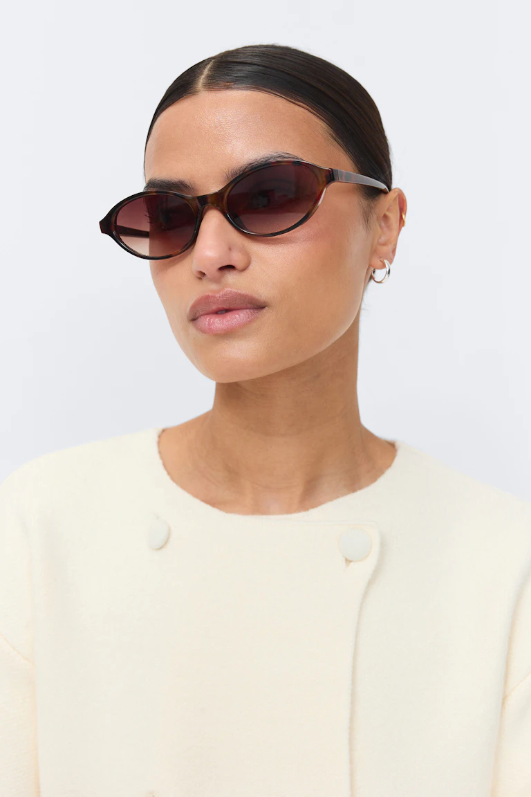 Tortoiseshell cat‑eye sunglasses with brown gradient lenses; small silver hoop earrings and a gold ear cuff; cream textured crew‑neck top with fabric‑covered shoulder buttons.