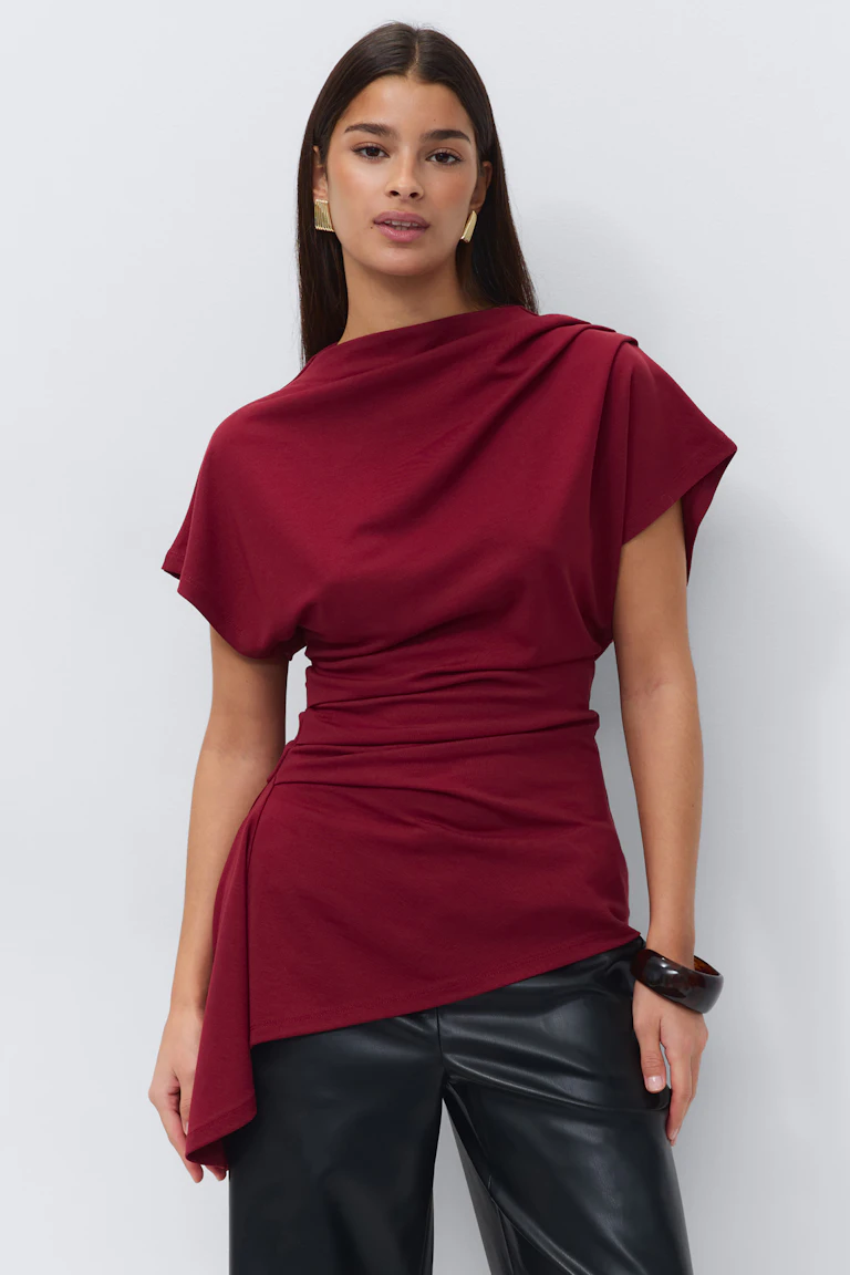 Top drapé asymétrique à manches courtes de couleur bordeaux avec taille froncée et ourlet angulaire, associé à un pantalon en cuir noir, des boucles d'oreilles rectangulaires dorées et un bracelet en écaille foncé.