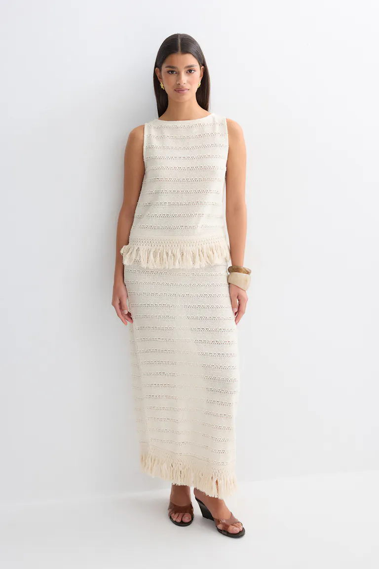Ivory sleeveless crochet-knit top with horizontal eyelet stripes and a fringe hem, paired with a matching ankle-length crochet-knit skirt with fringe trim; styled with brown open-toe heeled sandals, gold earrings and chunky beige bangles.