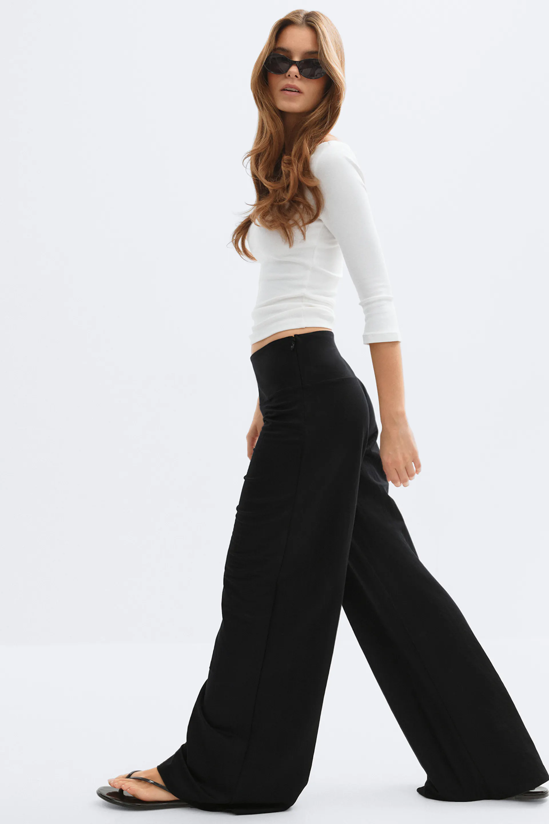 Weißes tailliertes Oberteil mit dreiviertel Ärmeln, schwarze Hose mit hoher Taille und weitem Bein, schwarze Zehensandalen und schwarze Sonnenbrille.