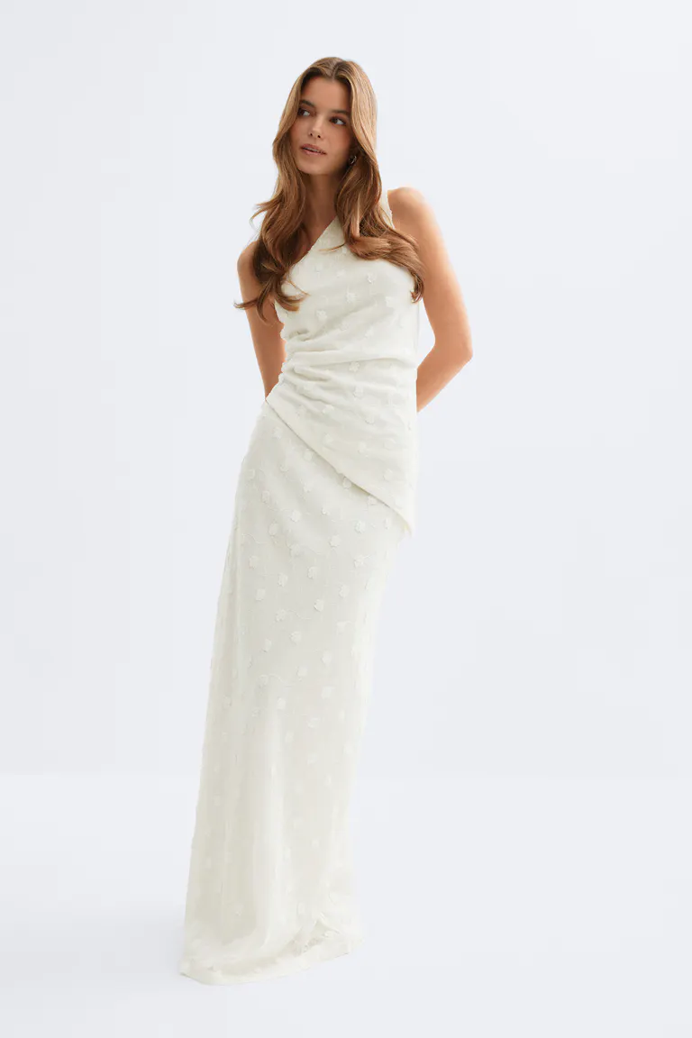Bodenlanges, elfenbeinfarbenes Kleid mit einer Schulter, dezenter floraler Jacquard-Textur, asymmetrisch drapierter Taille und schmaler Silhouette.