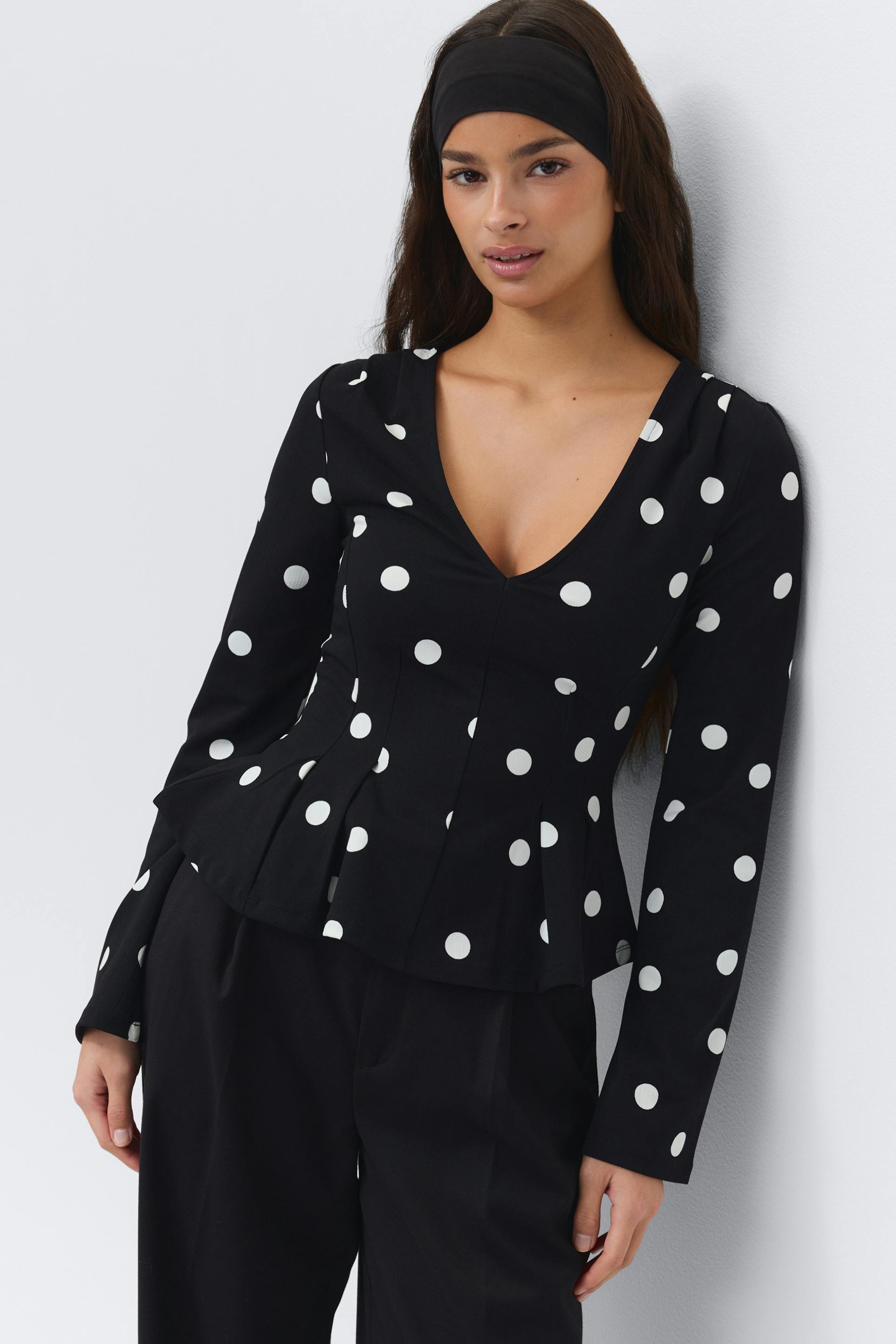 Black long-sleeve V-neck peplum blouse with white polka dots, styled with black tailored trousers and a wide black headband.