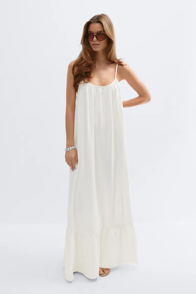 Floor-length ivory spaghetti-strap maxi dress with a gathered scoop neckline and ruffled hem, styled with oversized brown sunglasses, a silver bracelet, and tan flat sandals.