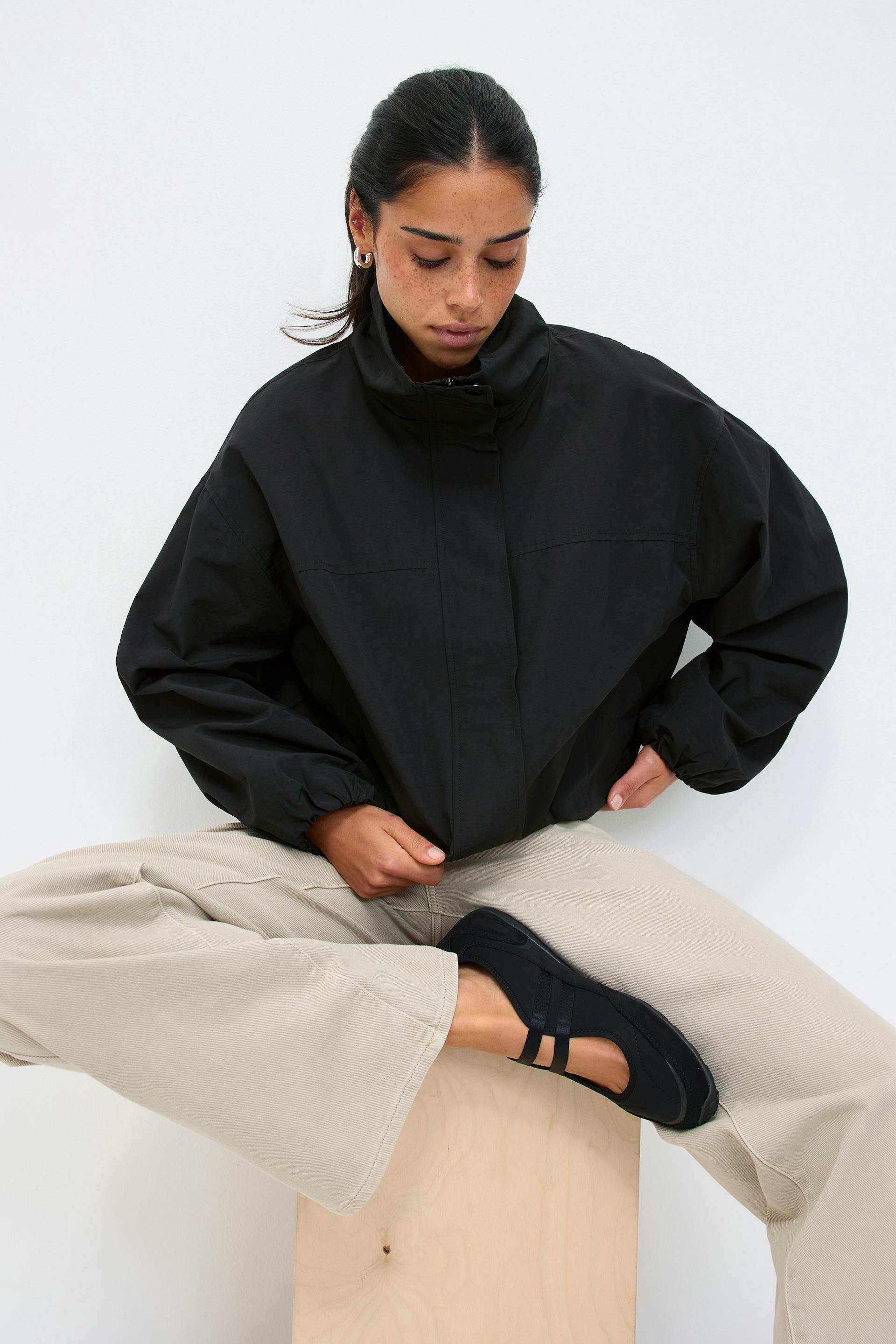 Schwarze übergroße Jacke mit hohem Kragen und Reißverschluss, beigefarbene Hose mit weitem Bein und schwarze Mary-Jane-Flats mit zwei Riemen.