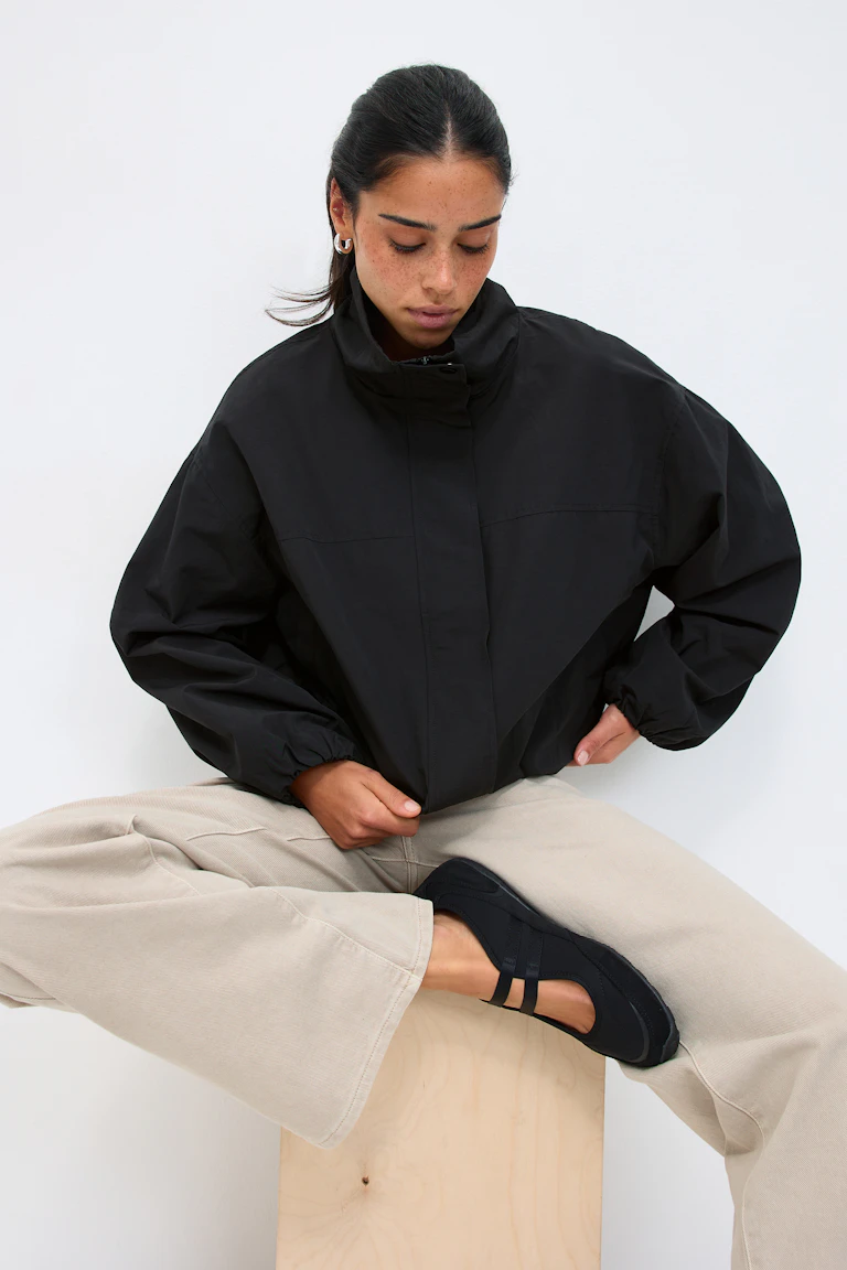 Schwarze übergroße Jacke mit hohem Kragen und Reißverschluss, beigefarbene Hose mit weitem Bein und schwarze Mary-Jane-Flats mit zwei Riemen.