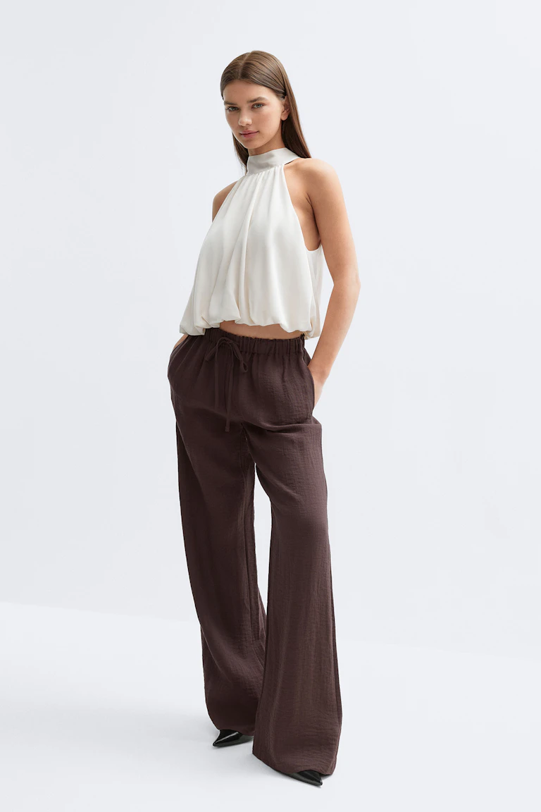 Witte halterblouse zonder mouwen met hoge hals en gerimpelde bolletjeszoom, gecombineerd met een donkerbruine broek met wijde pijpen en zwarte schoenen met spitse tenen.
