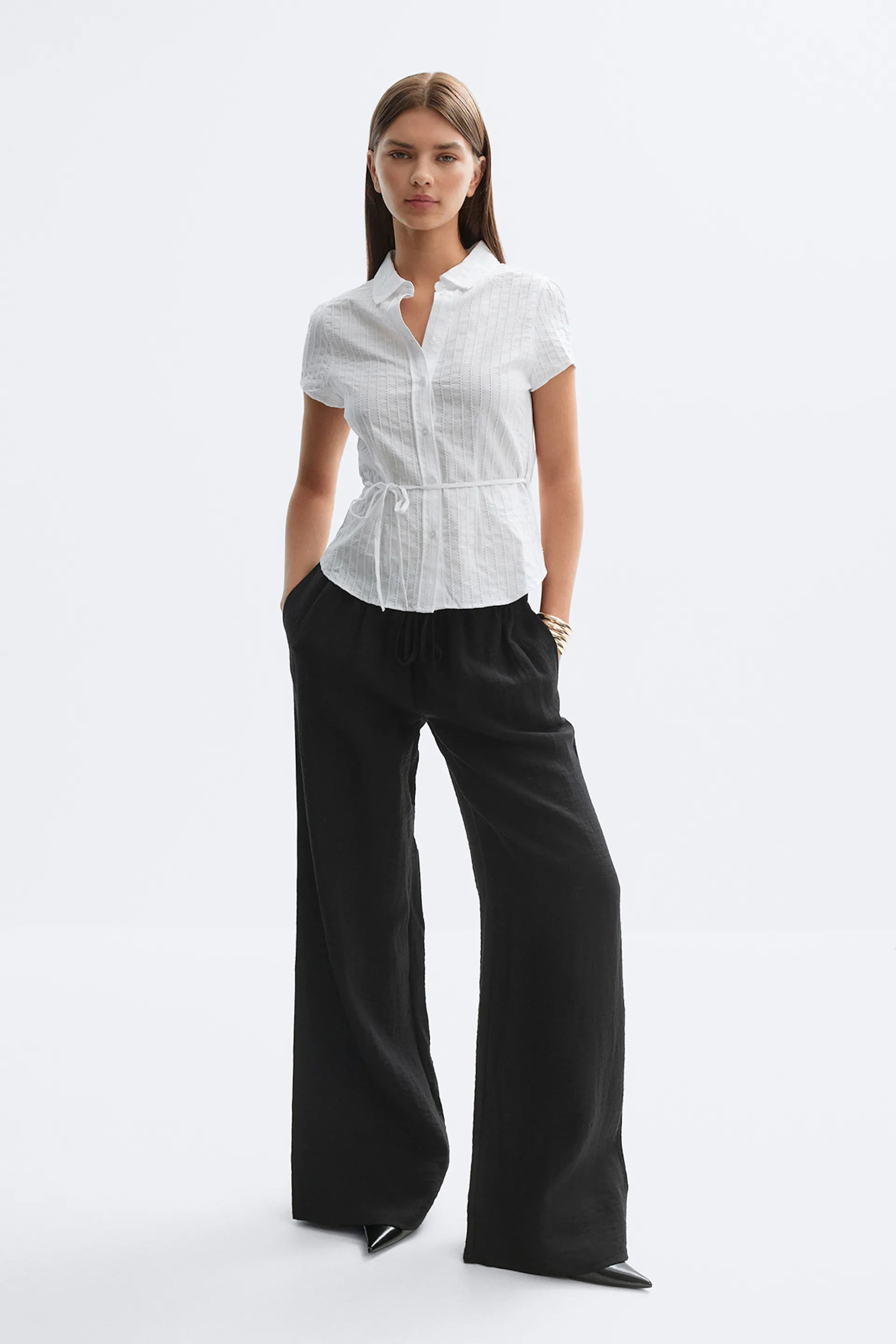 Weiße, kurzärmelige Button-Down-Bluse mit kleinem Kragen und Bindeband in der Taille, kombiniert mit einer schwarzen, hoch taillierten Hose mit weitem Bein, spitzen schwarzen Schuhen und goldenen Armreifen.
