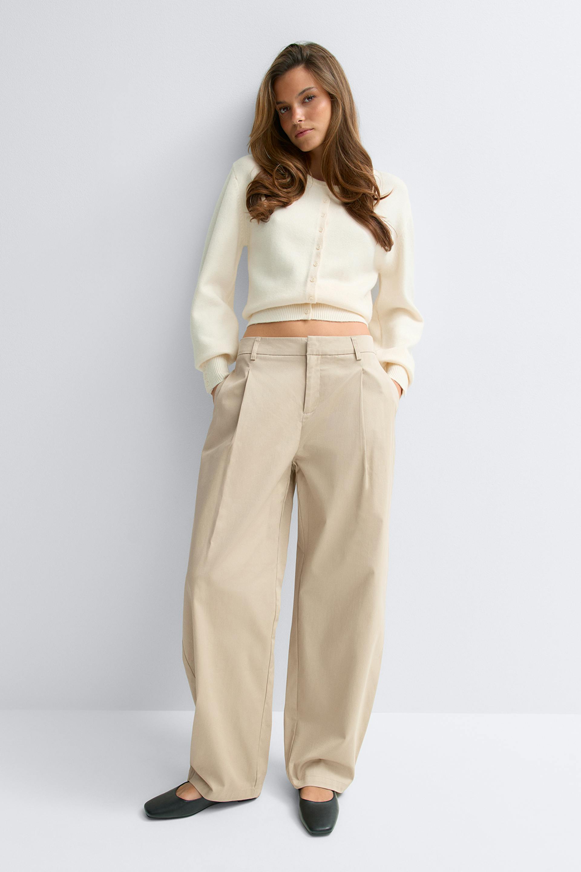 Cremefarbene, geknöpfte Strickjacke mit gerippten Bündchen und Saum, kombiniert mit einer beigefarbenen, weit geschnittenen Hose mit hoher Taille und schwarzen flachen Schuhen.