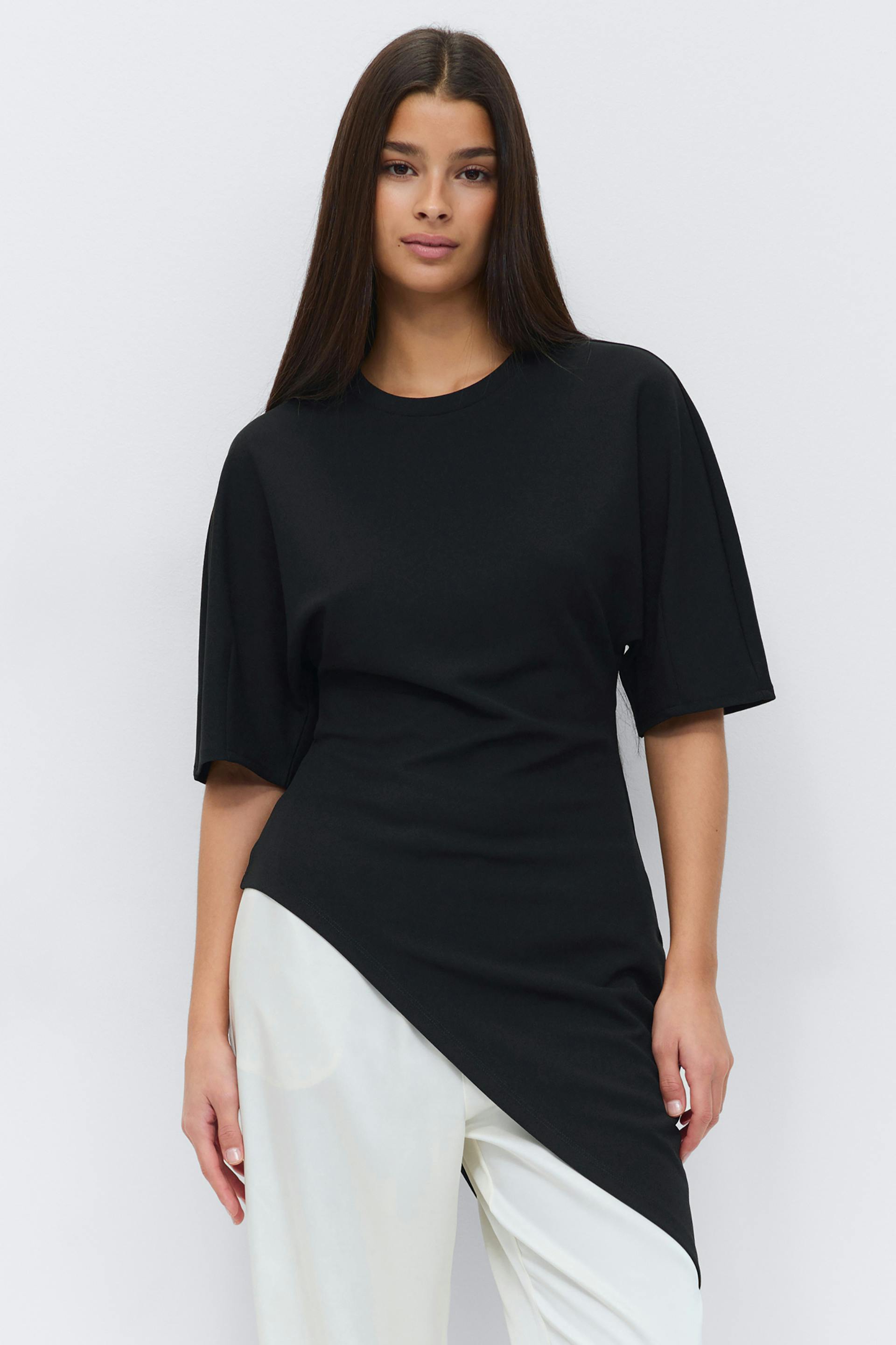 Black crew-neck tunic with elbow-length sleeves and an asymmetric diagonal hem, paired with white wide-leg trousers.