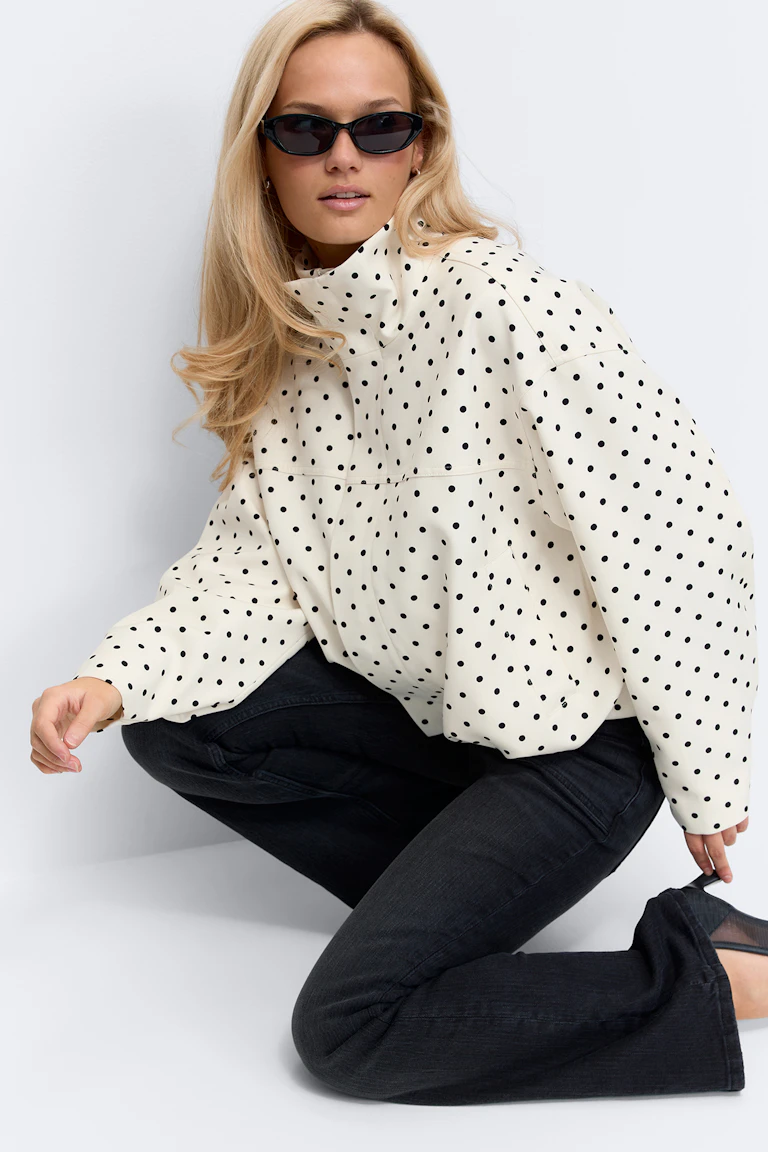Cremefarvet oversize jakke med høj hals, sorte prikker og lange ærmer, stylet med sorte slim jeans og sorte cat-eye solbriller.
