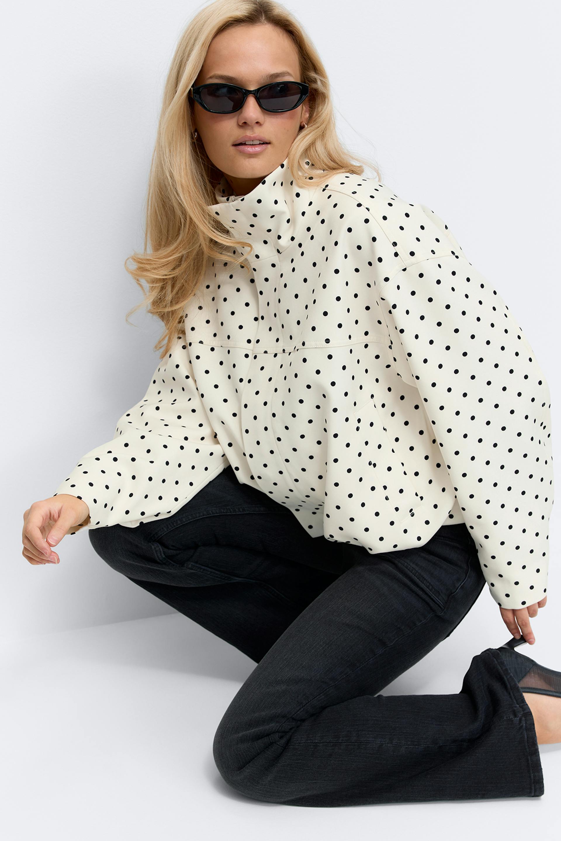 Cream high‑neck oversized jacket with black polka dots and long sleeves, styled with black slim jeans and black cat‑eye sunglasses.