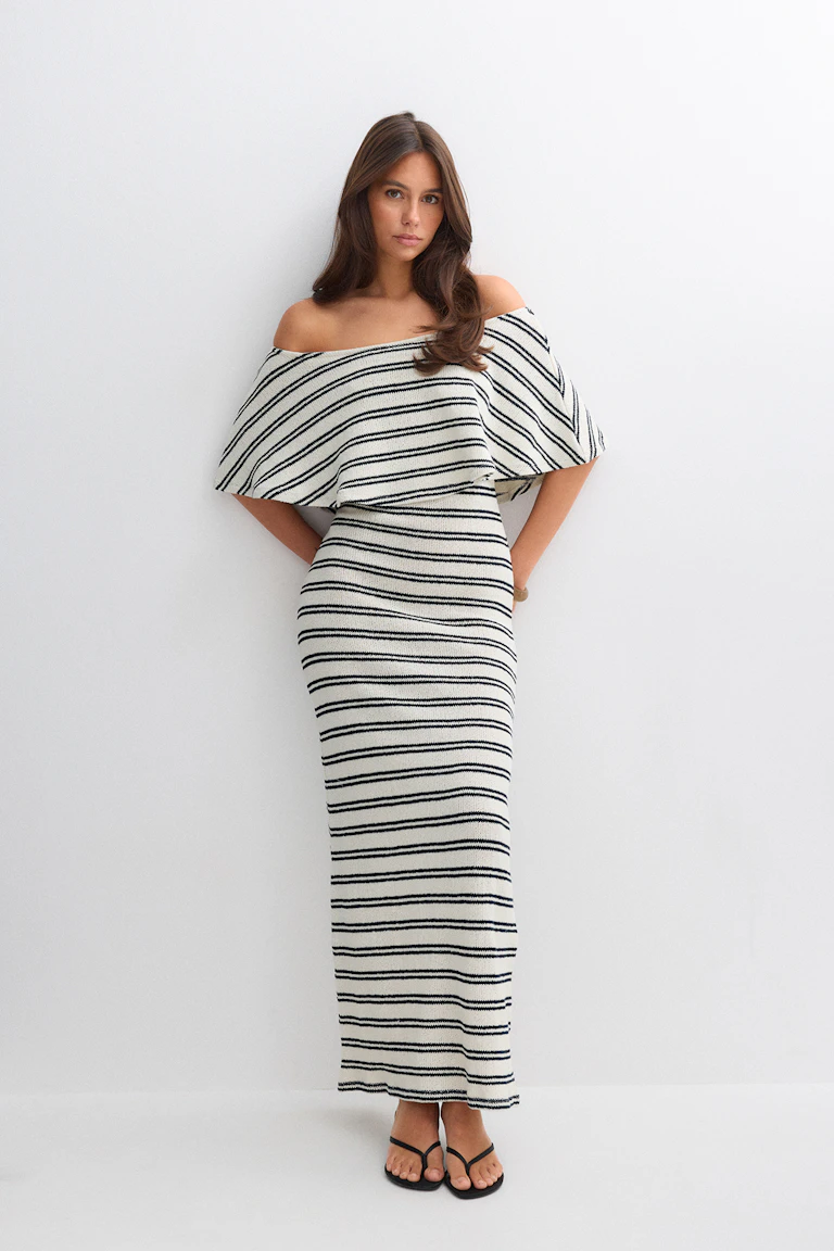 Cremefarbenes schulterfreies Strick-Maxikleid mit schmalen schwarzen Querstreifen, taillierter Silhouette und Capelet-Overlay; kombiniert mit schwarzen, dünnen Riemchensandalen.