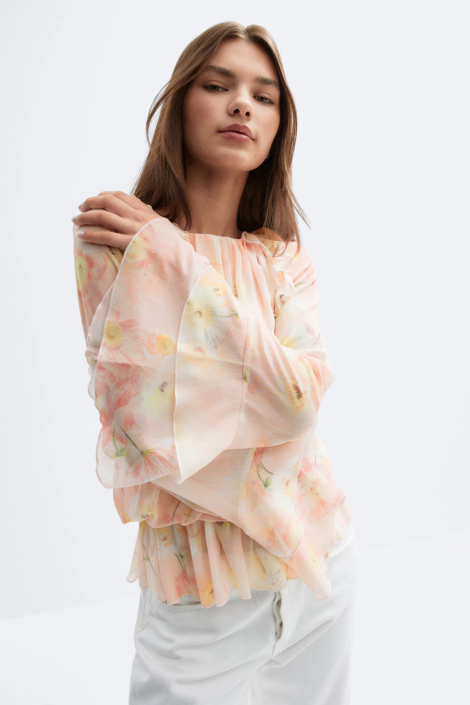 Durchsichtige, pastellfarbene Chiffonbluse mit gerafftem Ausschnitt, voluminösen Glockenärmeln und gerüschtem Schößchensaum, kombiniert mit einer weißen Hose mit hoher Taille.