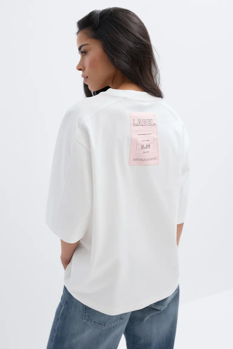 Weißes Oversized-T-Shirt mit ellbogenlangen Ärmeln und einem rosafarbenen, rechteckigen Etikett auf dem oberen Rücken, kombiniert mit einer lässigen Bluejeans.