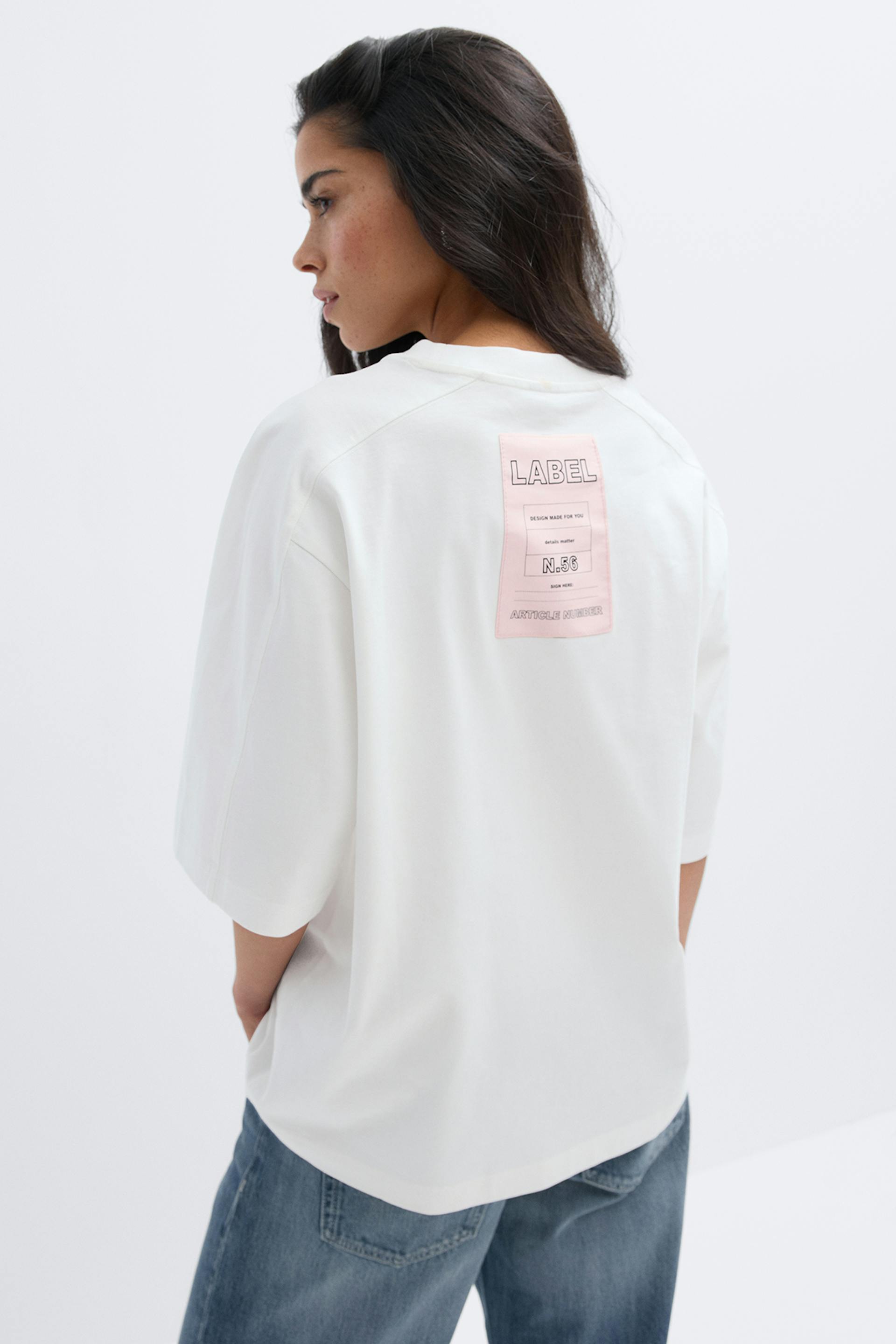 Valkoinen ylisuuri T-paita, jossa on kyynärpäiden pituiset hihat ja vaaleanpunainen suorakulmainen painettu etiketti selän yläosassa, yhdistettynä rennosti istuviin sinisiin farkkuihin.