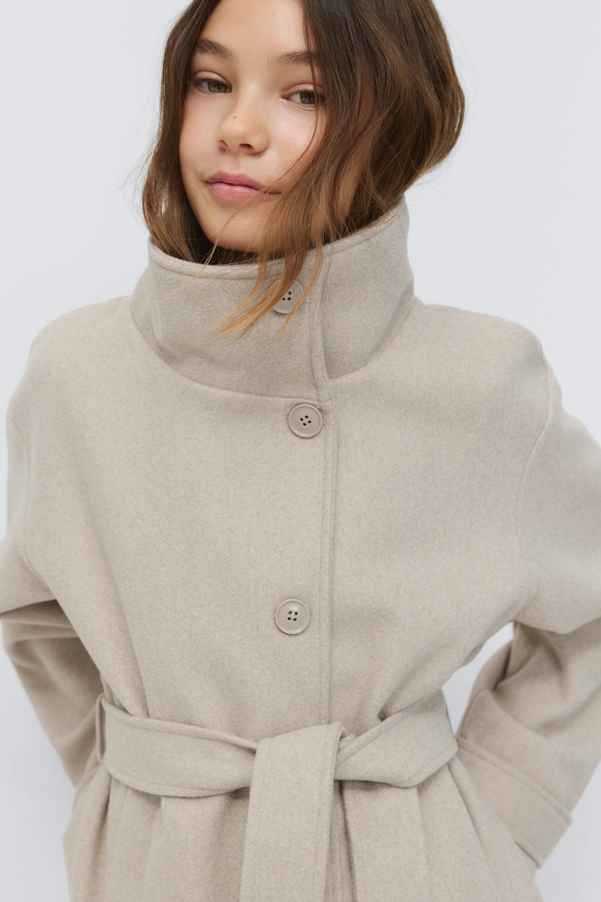 Ljusbeige ullrock med hög trattkrage, knappstängning framtill och midjebälte med självknytande band.