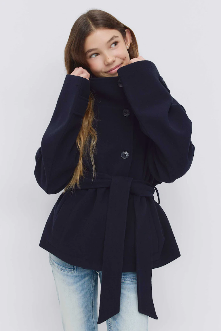 Marineblauer Wollmantel mit Gürtel, hohem Trichterkragen, übergroßen langen Ärmeln und Knopfverschluss vorne, kombiniert mit hellen Denim-Jeans.