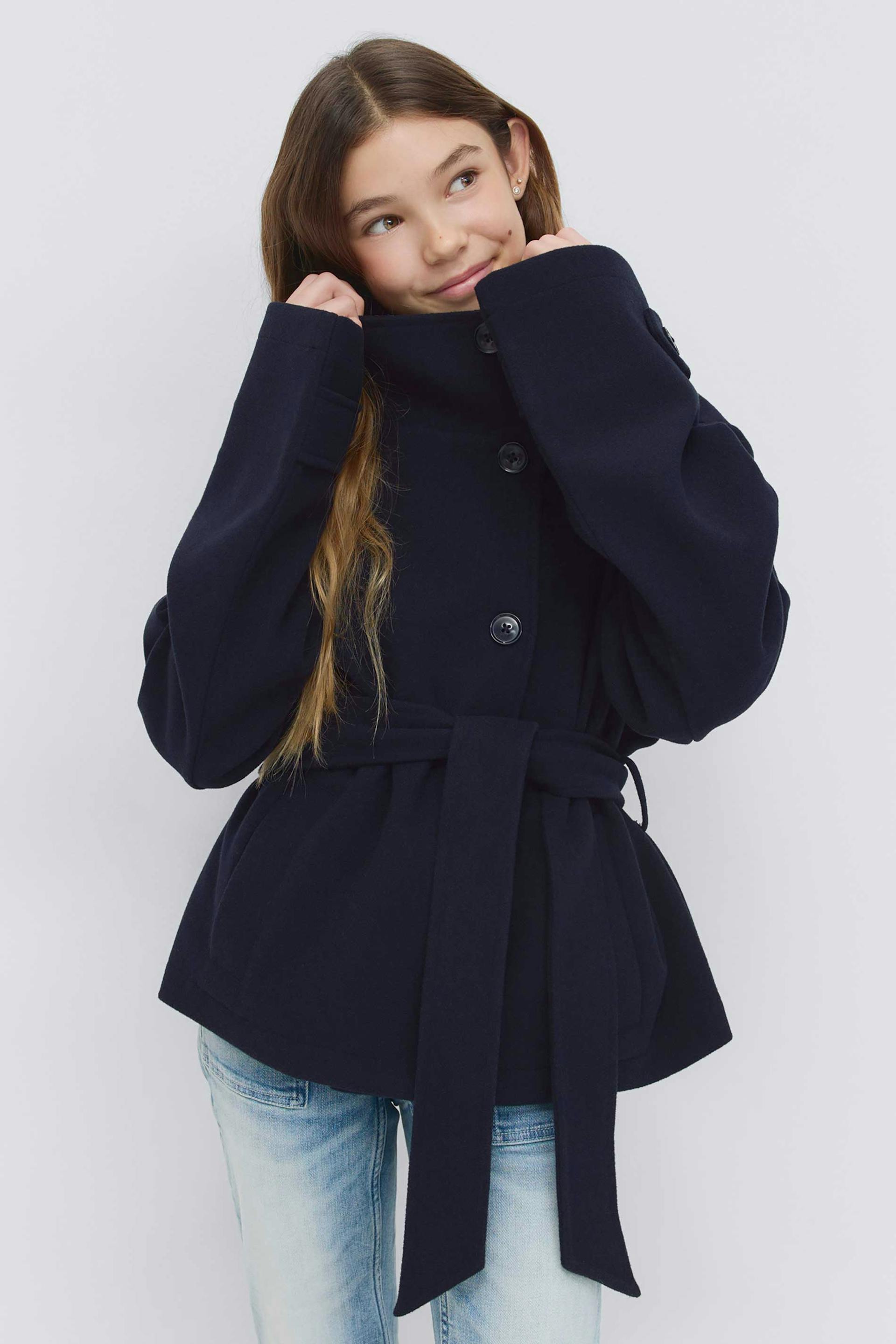 Marinblå, bältad ullkappa med hög trattkrage, överdimensionerade långa ärmar och knappstängning fram, kombinerad med ljusa jeans.