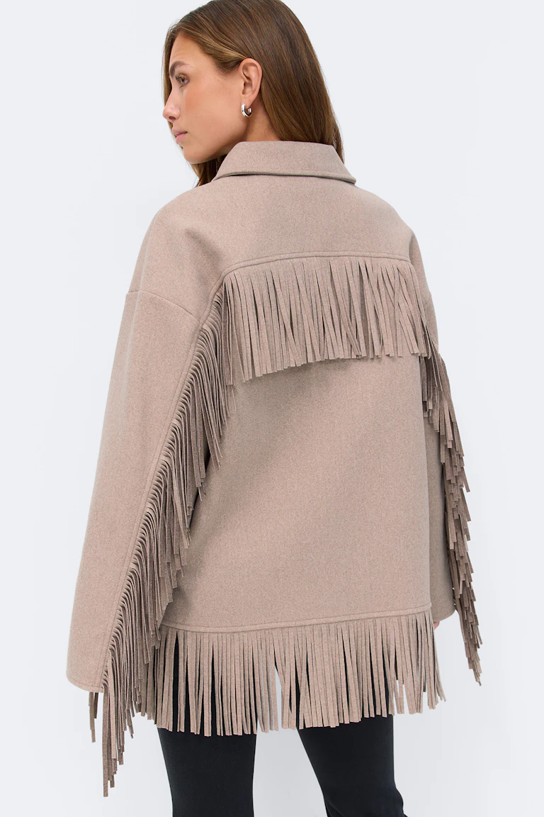 Taupefarbene Fransenjacke aus einer Wollmischung mit gekerbtem Kragen und langen Ärmeln, mit Fransenbesatz am Rücken, an den Ärmeln und am Saum; kombiniert mit einer schwarzen schmalen Hose.