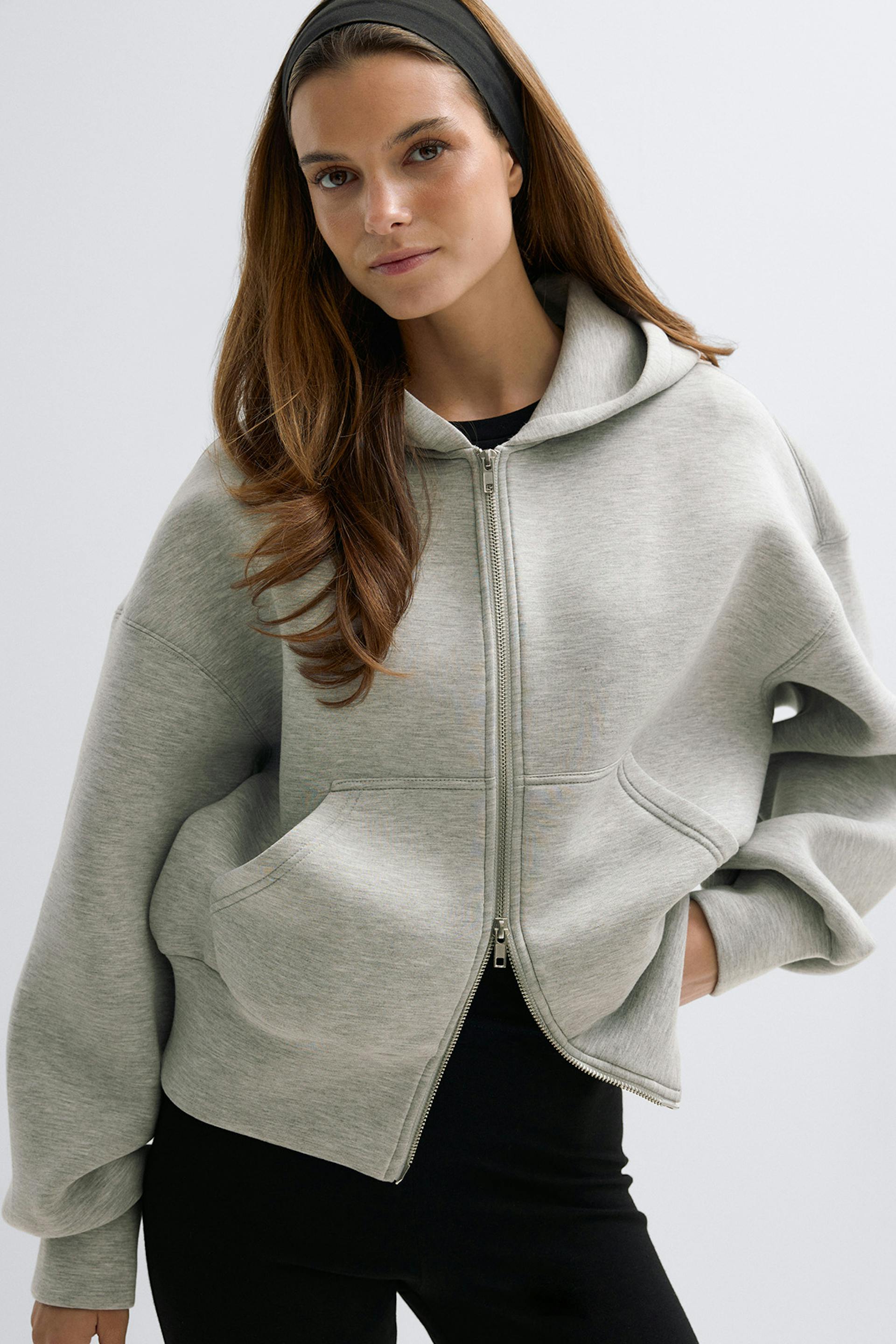 Sweat à capuche zippé surdimensionné gris clair avec capuche, poches kangourou sur le devant et fermeture éclair argentée, associé à un pantalon ajusté noir et à un bandeau noir.