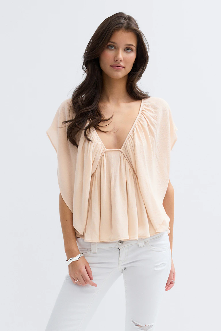 Pale-peach plisserad djup V-ringad blus med draperade cap-ärmar, kombinerad med vita slitna skinny jeans och ett silverfärgat armband med manschett.