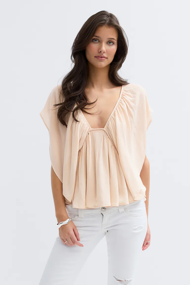 Pale-peach plisserad djup V-ringad blus med draperade cap-ärmar, kombinerad med vita slitna skinny jeans och ett silverfärgat armband med manschett.