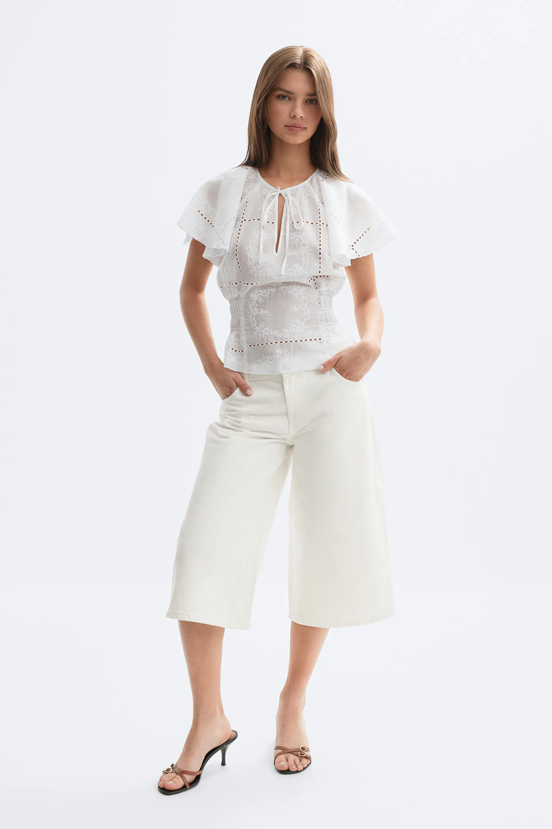 Weiße bestickte Ösenbluse mit Flatterärmeln, Schlüssellochausschnitt zum Binden und gesmokter Taille, kombiniert mit einer cremefarbenen Hose mit weitem Bein und braunen Pantoletten mit Riemchenabsätzen.