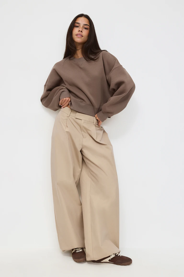 Sweat oversize marron à col rond et épaules tombantes ; pantalon beige à taille haute et à jambes larges ; baskets à lacets marron et blanches.