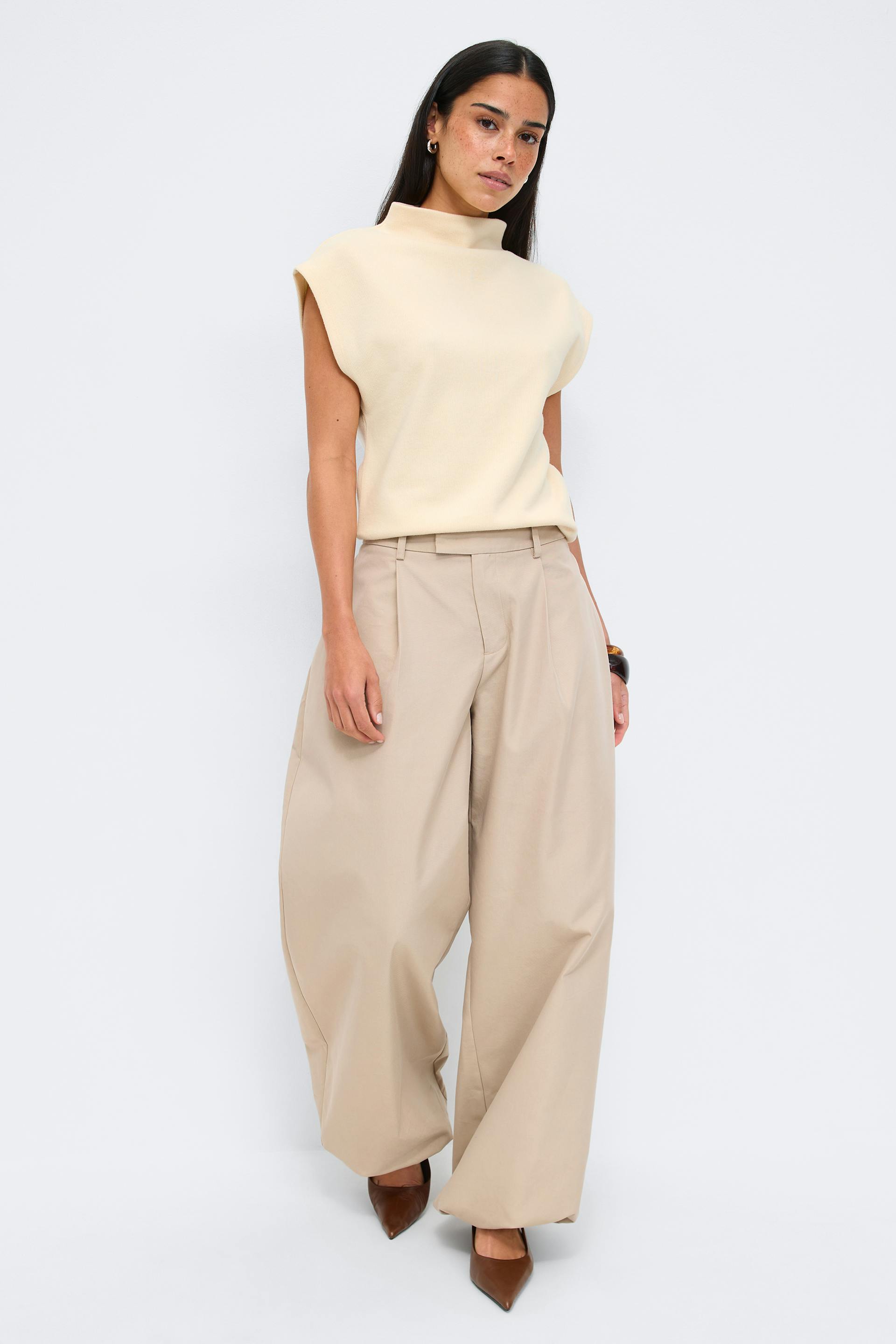 Krämfärgad ärmlös topp med mockahals kombinerad med beige vida plisserade byxor med hög midja och bruna spetsiga klackar.