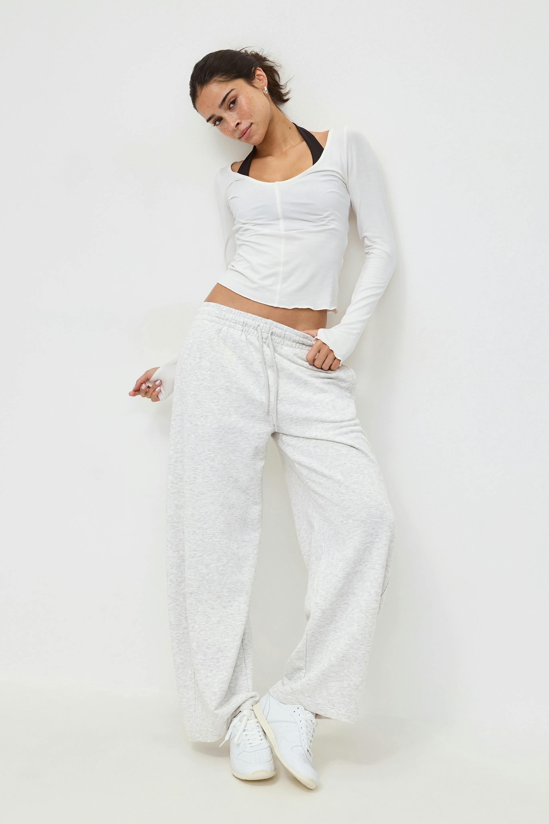 White long-sleeve fitted scoop-neck top layered over a black sports bra, light heather-gray wide-leg drawstring sweatpants, and white low-top sneakers.