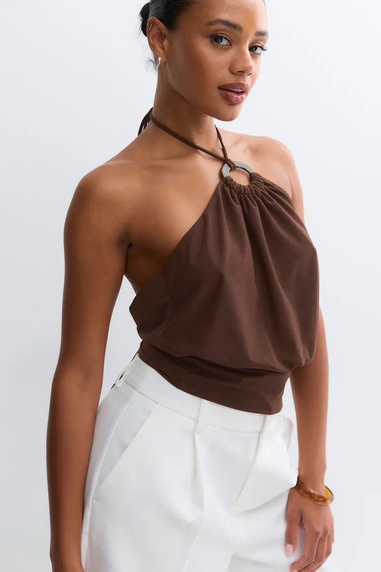 Braunes Neckholder-Top mit Metallring-Detail und gerafftem Ausschnitt, das mit einem taillierten Saum abschließt, gepaart mit einer weißen, hoch taillierten Hose.