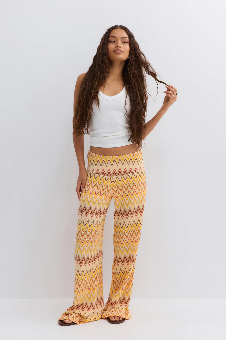 White sleeveless V-neck cropped tank top paired with high-waisted, wide-leg knit trousers in a yellow, orange and brown zigzag pattern, and brown flat sandals.