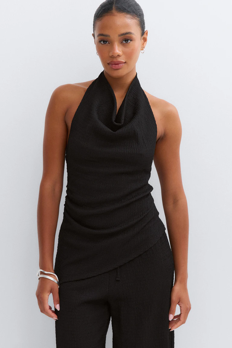 Débardeur sans manches texturé noir avec encolure vache drapée et ourlet asymétrique, associé à un pantalon texturé noir assorti ; coiffé de petites boucles d'oreilles argentées et de bracelets argentés superposés.