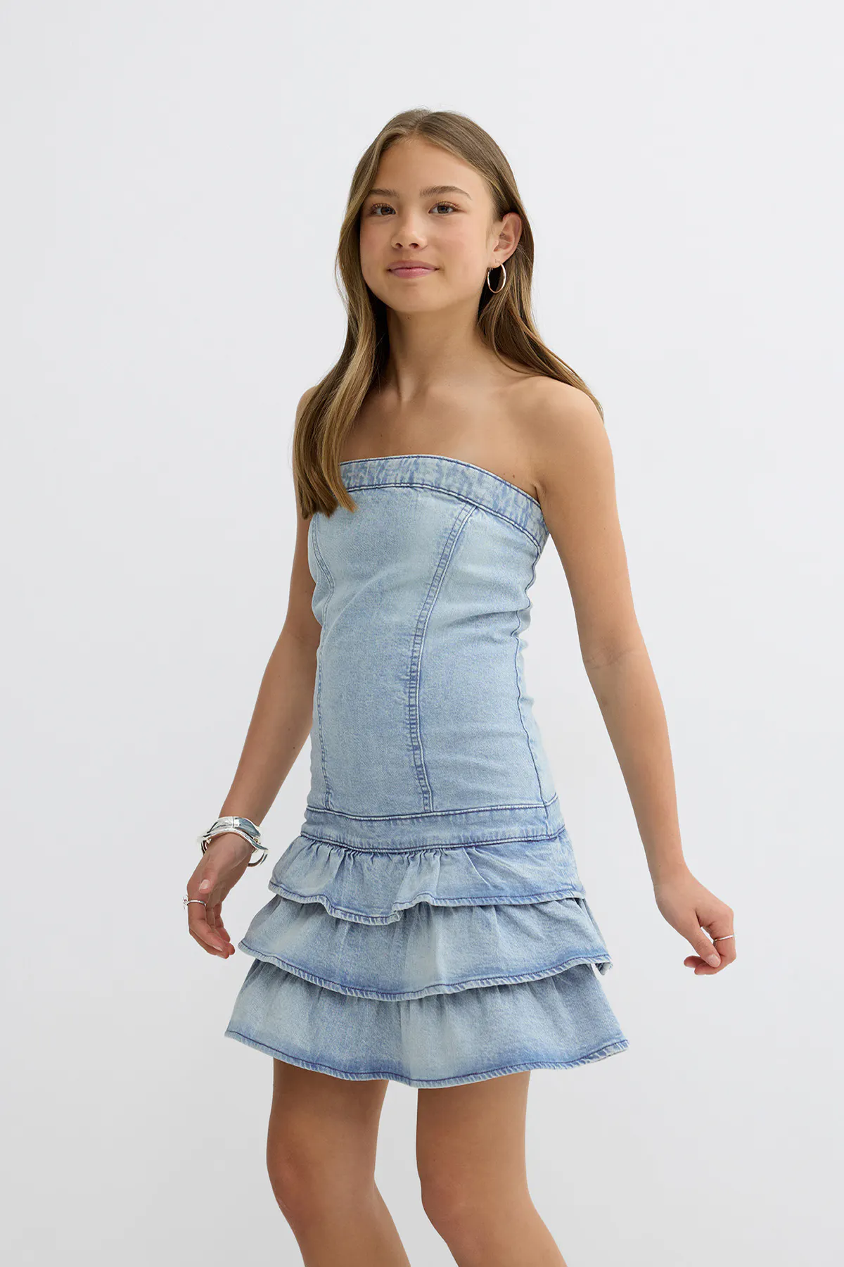 Mini robe bustier en denim délavé avec un corsage ajusté et une jupe à volants, associée à des boucles d'oreilles argentées, un bracelet manchette argenté et des bagues.