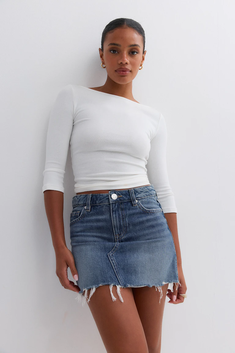 White fitted asymmetrical boat-neck top with three-quarter sleeves, paired with a high‑waist blue denim mini skirt with frayed hem; gold hoop earrings.