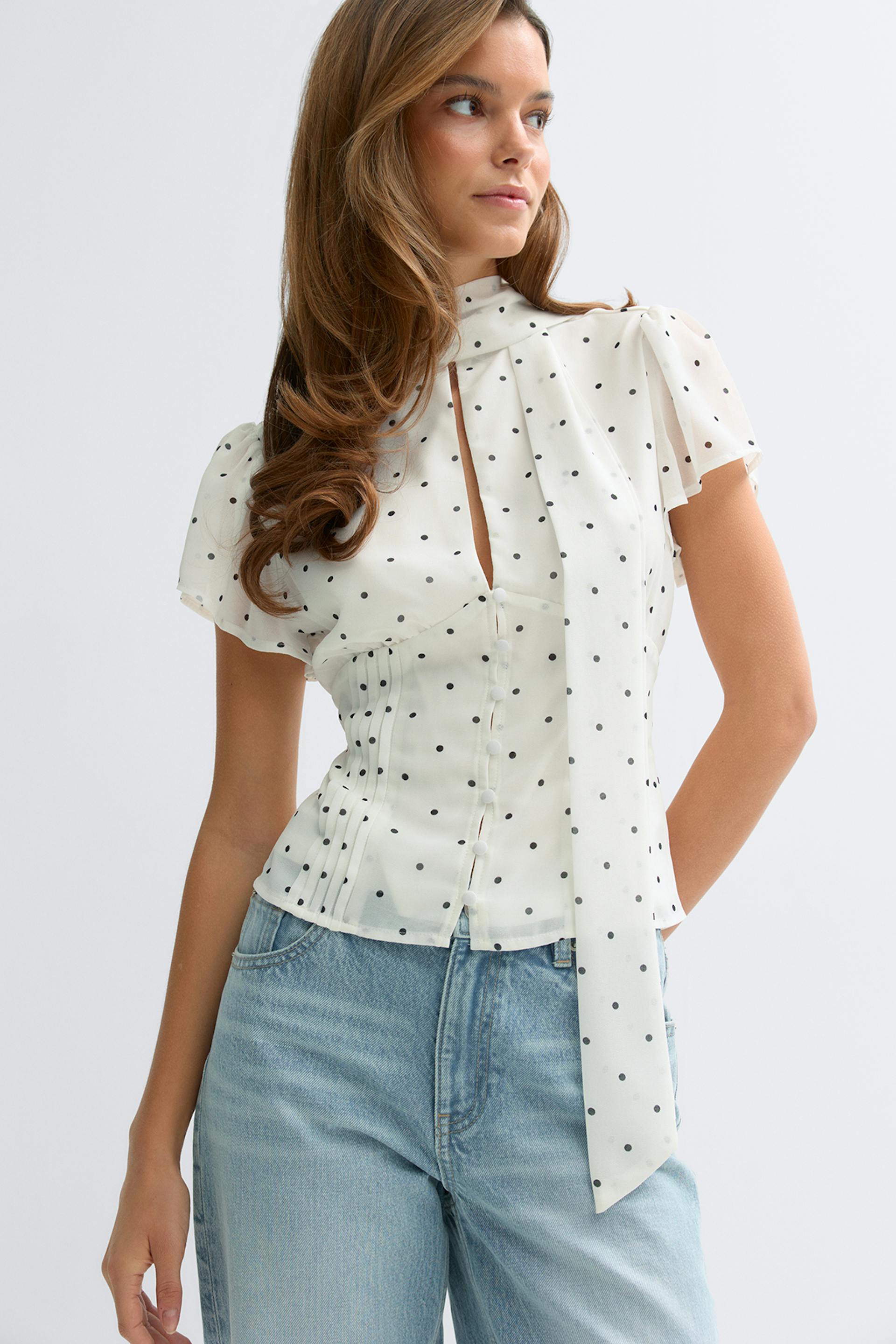 Weiße, durchsichtige, kurzärmelige Polka-Dot-Bluse mit schwarzen Punkten - hoher Halsausschnitt mit langem Schal zum Binden, Schlüsselloch auf der Vorderseite und Mieder mit verdeckten Knöpfen - kombiniert mit hellen, hoch sitzenden Jeans.