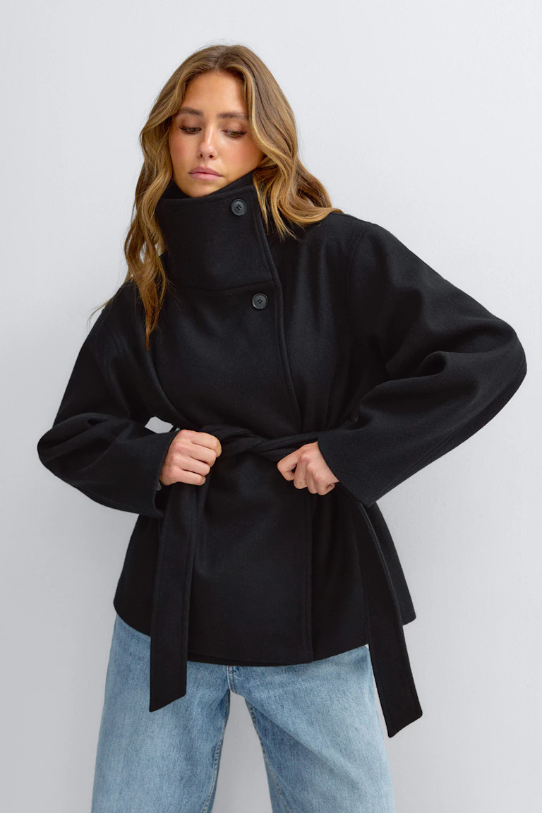 Schwarzer Wollmantel mit hohem Kragen und übergroßem Knopfverschluss, kombiniert mit einer Jeans mit geradem Bein und heller Waschung.