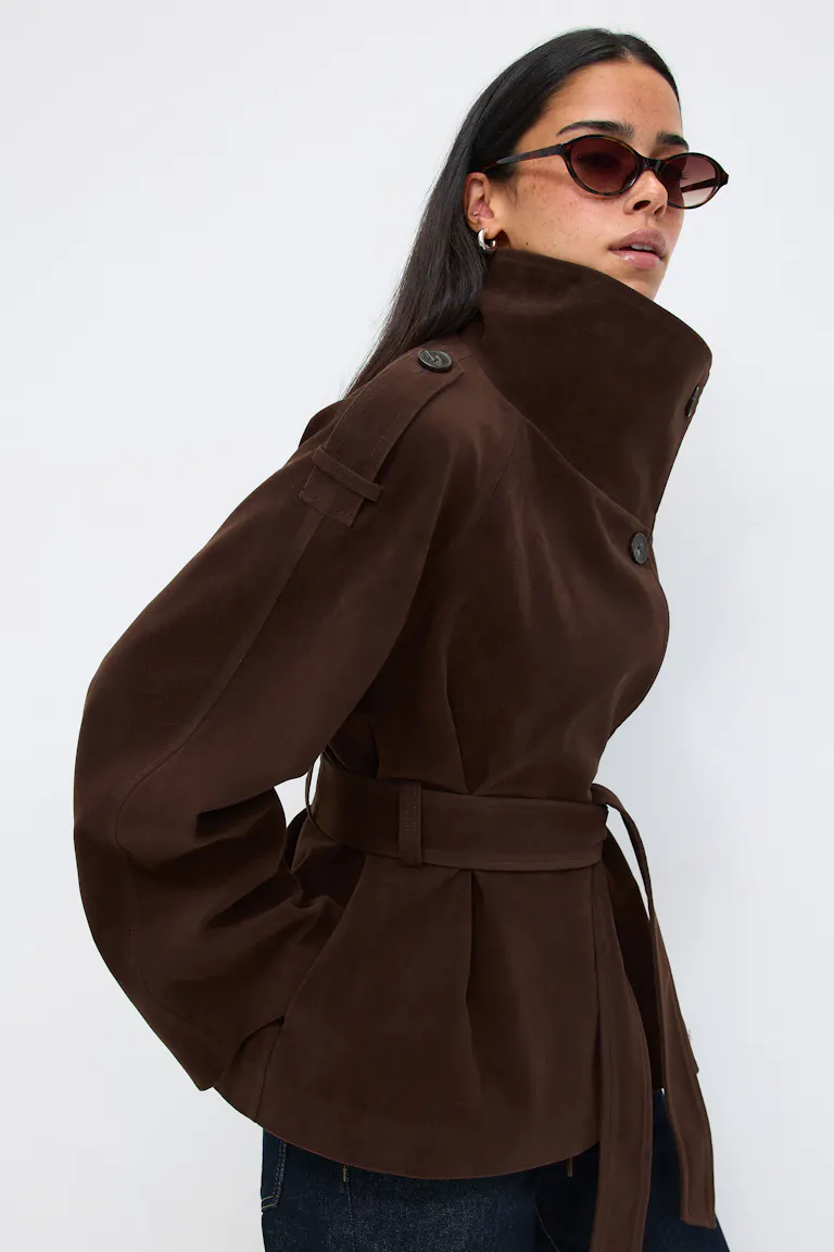 Dark brown suede belted jacket with high funnel collar, buttoned epaulettes and voluminous sleeves, styled with tortoiseshell cat‑eye sunglasses and dark denim jeans.